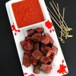 Overhead view of Halloween steak bites on a white platter with tomato garlic dip, blood splatter decorations and bamboo skewers