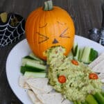 Carved pumpkin throwing up spicy guacamole onto a platter with low carb chips, cucumber and celery sticks