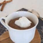 Low carb chocolate mug cake in a white cup topped with whipped cream, with cinnamon sticks behind