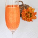 Orange glitter Halloween cocktail in a champagne flute with a small pumpkin and orange flower behind it