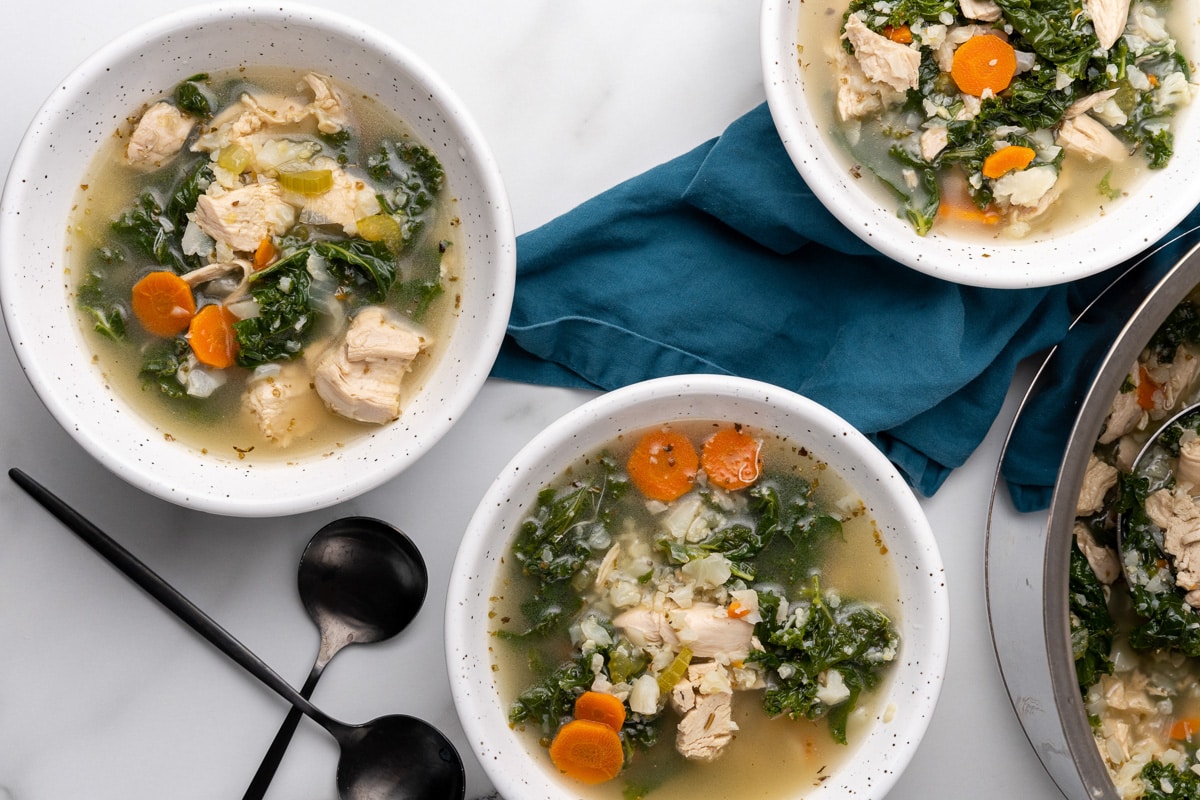 Two bowls of chicken kale soup with black spoons next to the pot on a marble counter