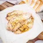 Two baked lemon pepper tilapia filets on a white square plate on a wooden board