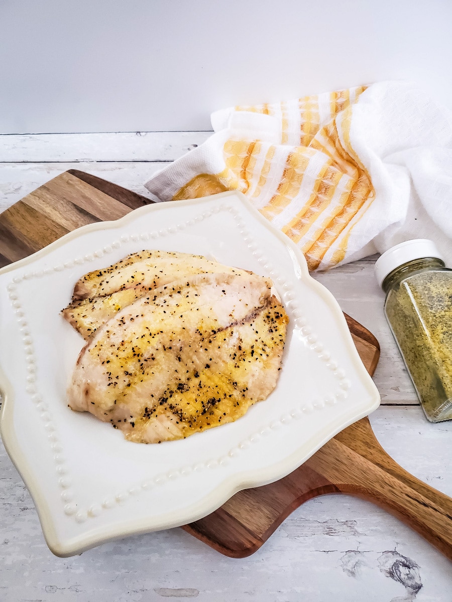 Lemon pepper tilapia on a white plate with a striped kitchen towel in the background