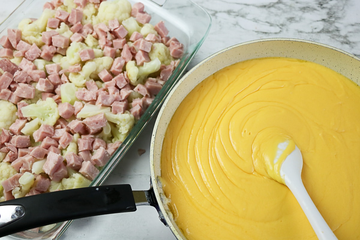 Cubed ham scattered over cauliflower in a baking dish next to a skillet of cheddar cream sauce