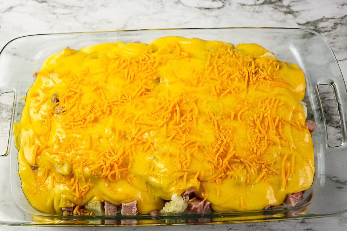 Ham cauliflower casserole covered in cheese sauce and shredded cheddar, ready to bake