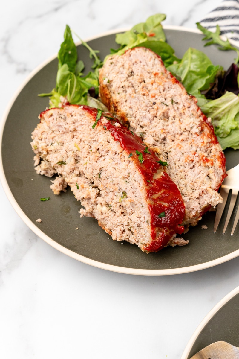 Two slices of turkey meatloaf served with mixed salad greens on a dark plate