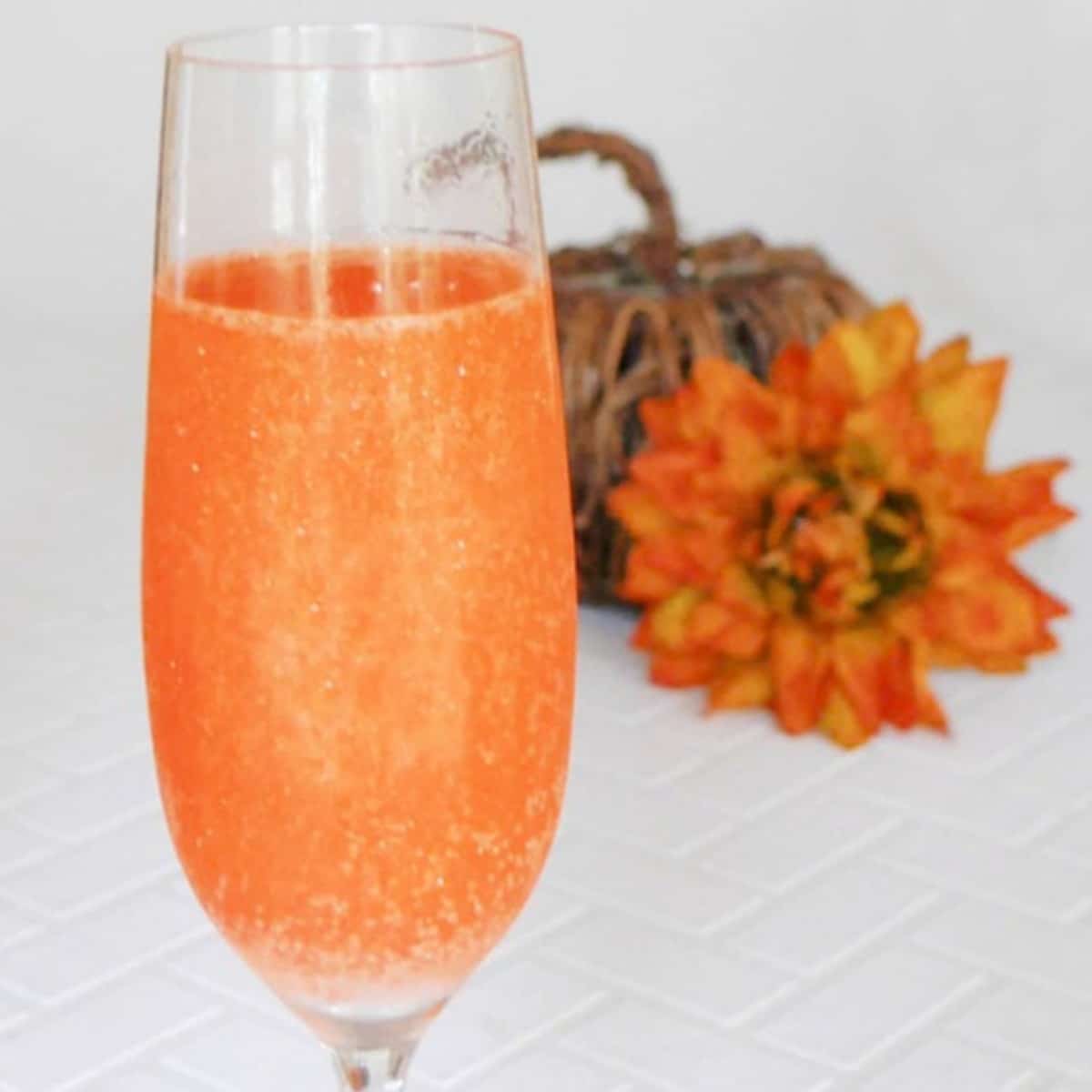 Orange glitter Halloween cocktail in a champagne flute with a small pumpkin and orange flower behind it