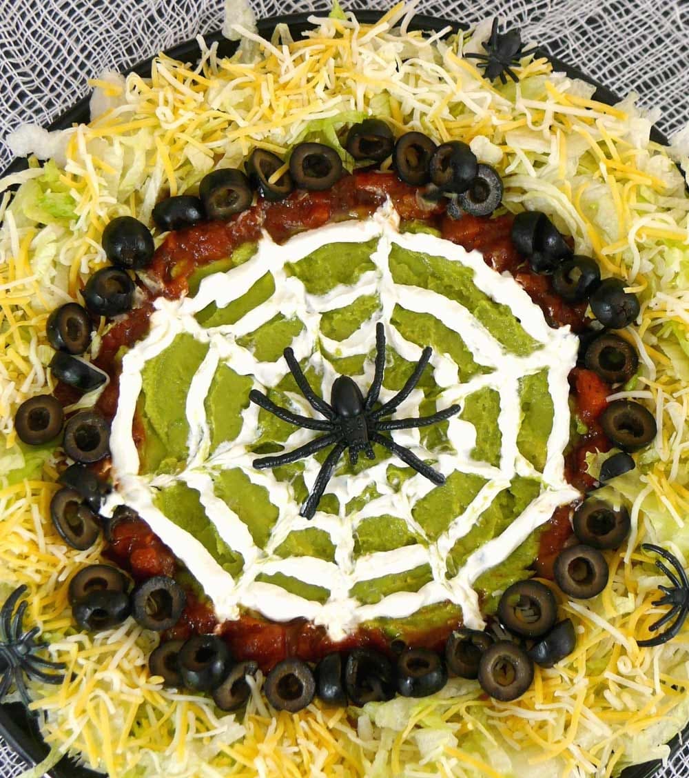 Overhead view of a Halloween spider web dip with a sour cream web piped over guacamole, ringed with salsa, black olives, lettuce and cheese