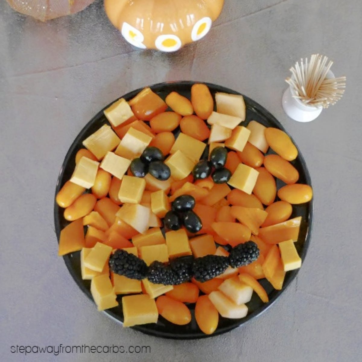 Keto Halloween snack board shaped like a jack-o'-lantern, with cheddar, orange peppers, cantaloupe and tomatoes and an olive and blackberry face