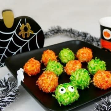 Low carb monster bites rolled in orange and green coconut on a black Halloween platter, one with candy eyes