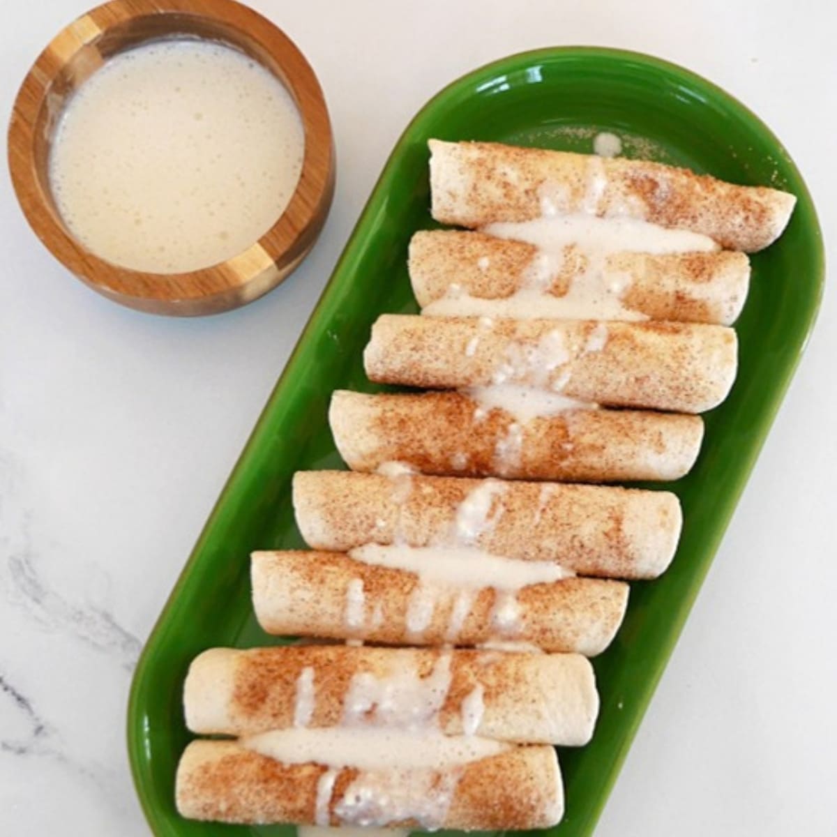 Low carb tortilla cinnamon roll-ups on a green platter with a bowl of cream cheese glaze