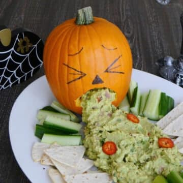 Carved pumpkin puking spicy guacamole onto a platter with low carb chips, cucumber and celery sticks