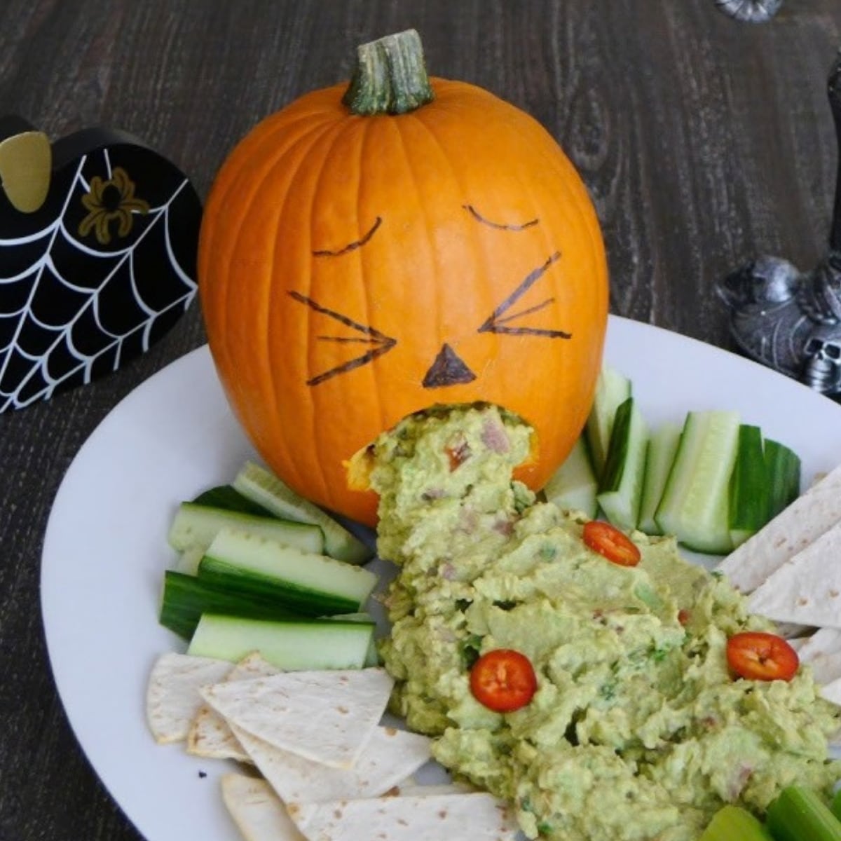 Carved pumpkin puking spicy guacamole onto a platter with low carb chips, cucumber and celery sticks