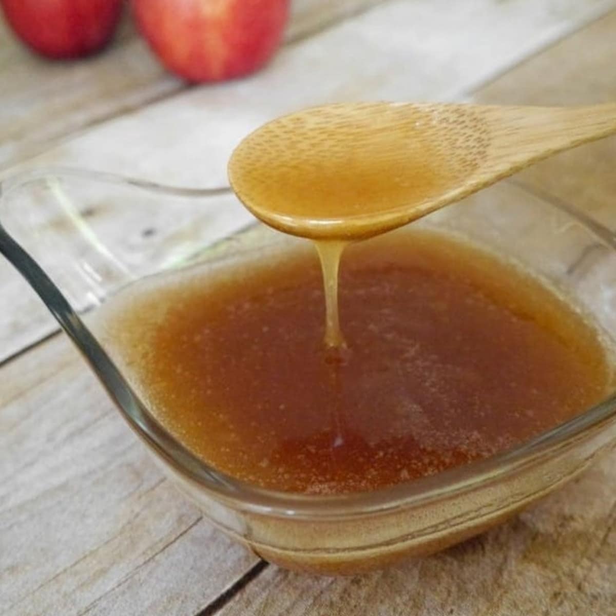 Low carb caramel apple sauce dripping from a wooden spoon into a glass bowl with red apples behind